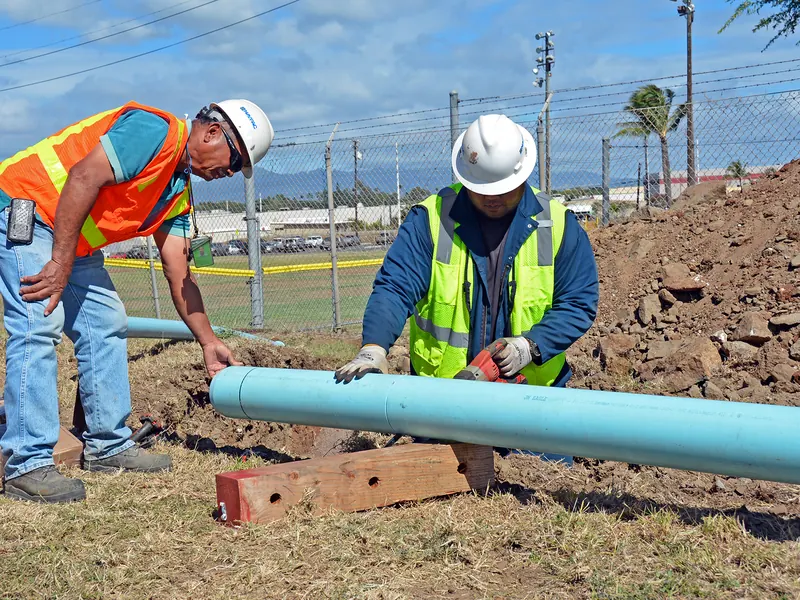 Two workers in hard hats and high-visibility vests fitting a water line pipe on site
